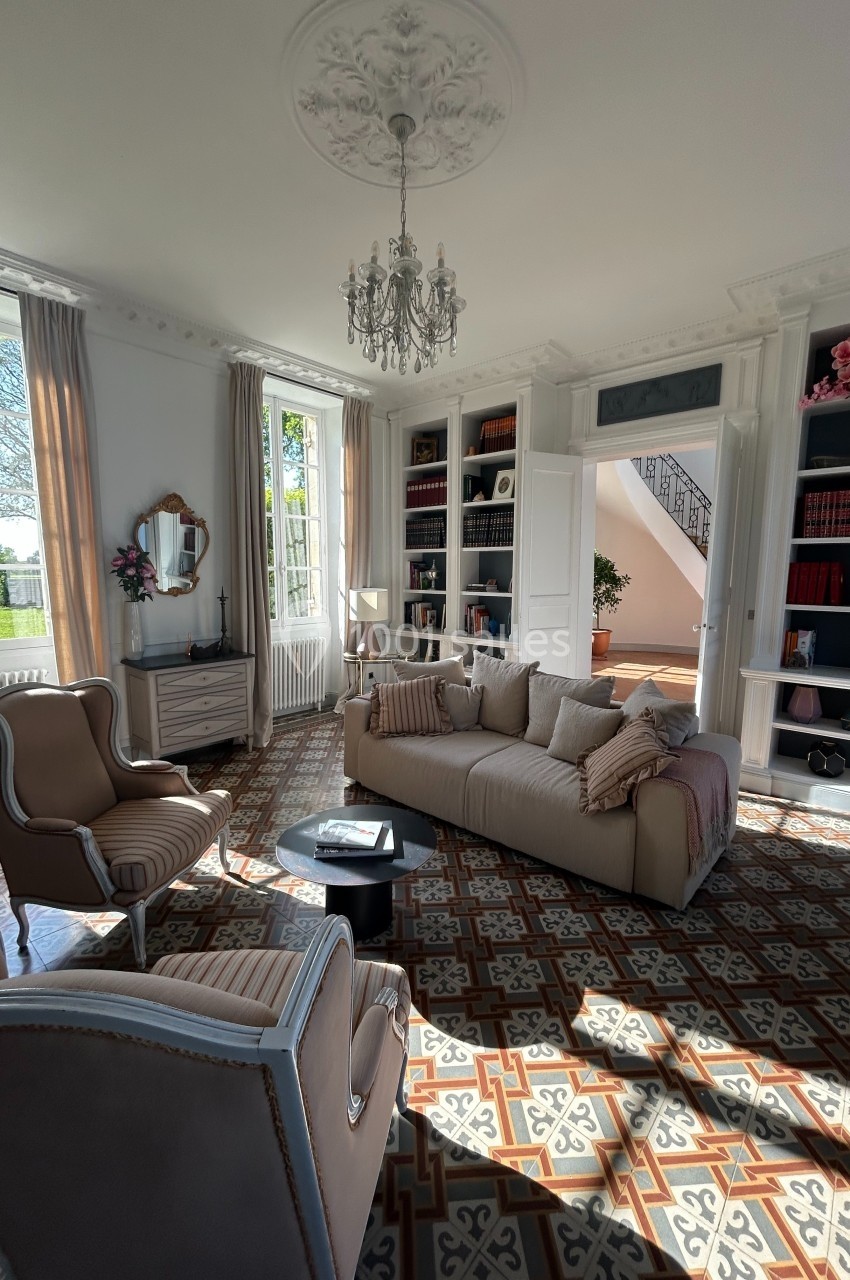 Salon lumineux avec canapé beige, fauteuils assortis, bibliothèque intégrée, lustre et carrelage à motifs.