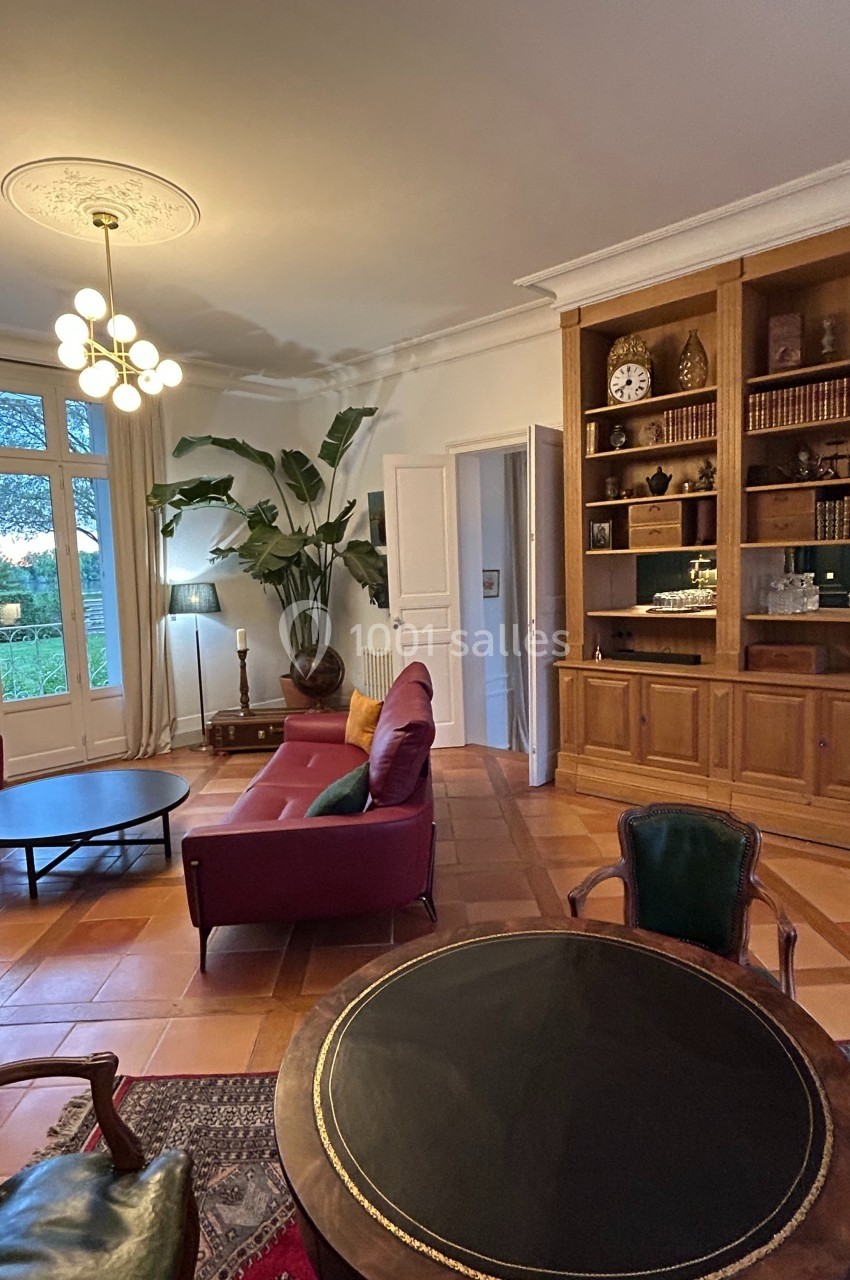 Salon lumineux avec mobilier classique, bibliothèque en bois, grandes fenêtres et vue sur un jardin verdoyant.