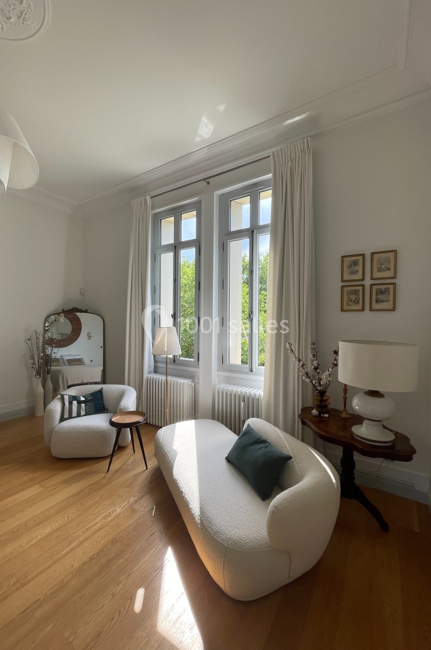Salon lumineux avec parquet, fauteuils modernes, table basse, miroir ancien et grande fenêtre donnant sur un jardin.