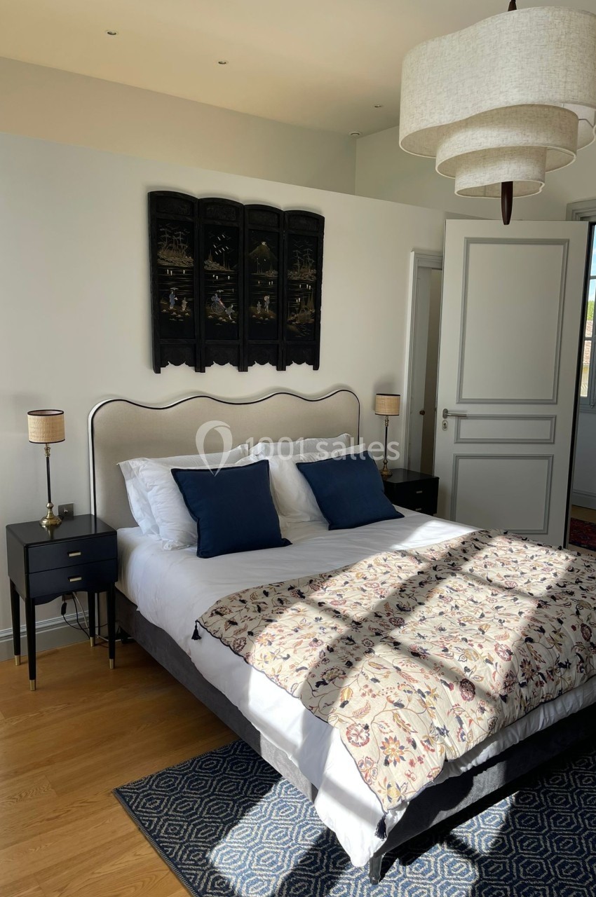 Lit double avec tête de lit beige, coussins bleus, lampes de chevet et couvre-lit à motifs dans une chambre lumineuse.