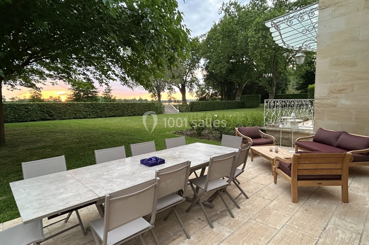 Terrasse avec table et chaises, vue sur un jardin verdoyant et un coucher de soleil à l'horizon.