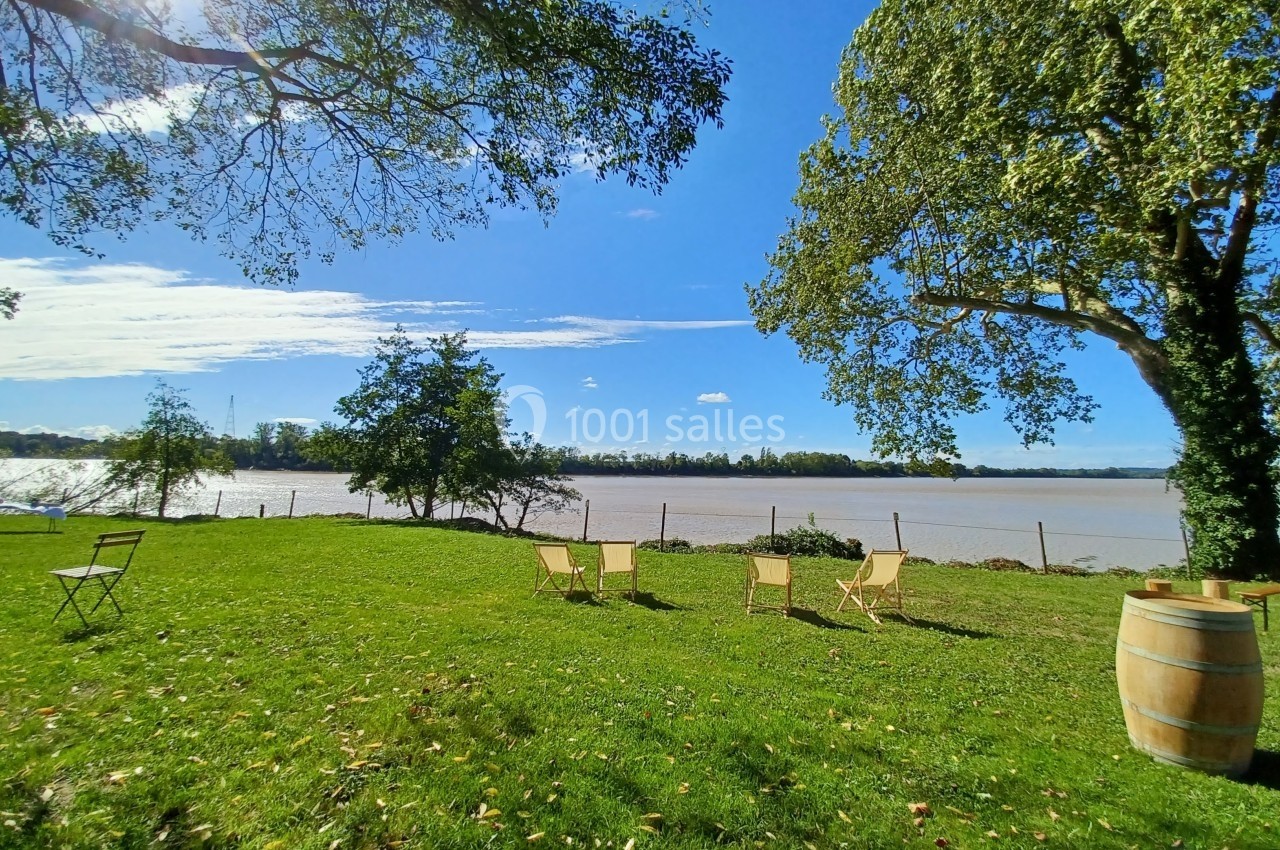 Chaises et table disposées sur une pelouse verdoyante près d'un fleuve, entourées d'arbres sous un ciel bleu.