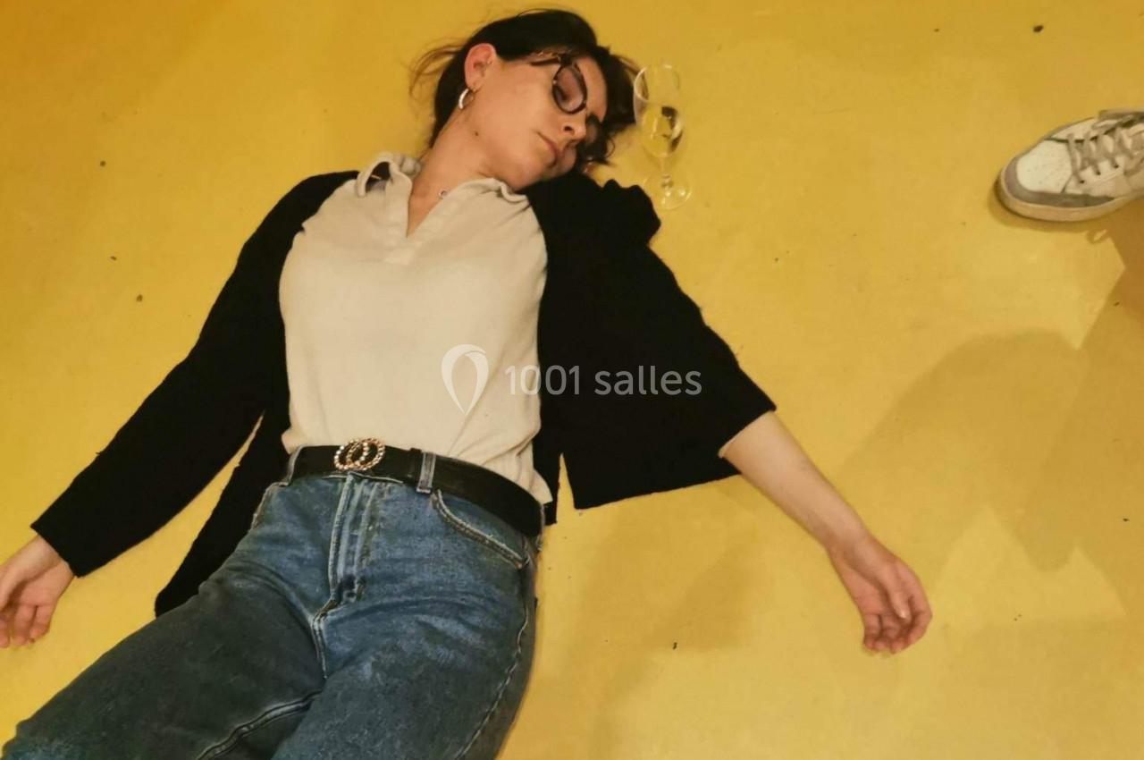 Femme allongée sur le sol jaune, lunettes tombées à côté d'elle, avec une chaussure visible en arrière-plan.