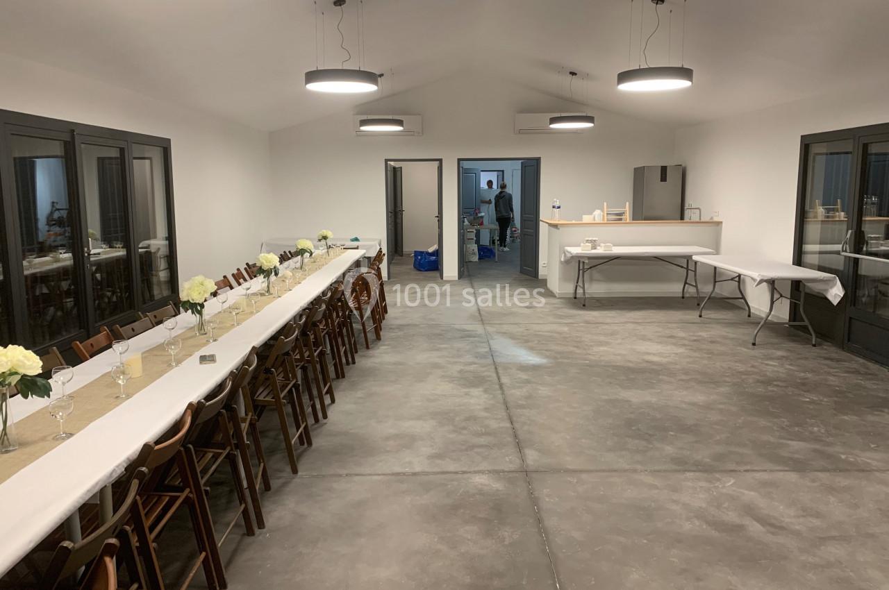Salle aménagée avec de longues tables dressées, des chaises en bois et un espace cuisine en arrière-plan.