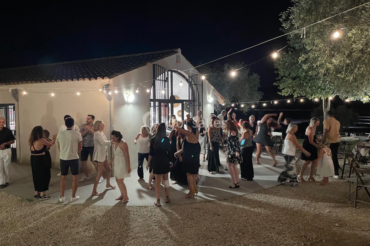 Groupe de personnes dansant et discutant en soirée en plein air, éclairée par des guirlandes lumineuses près d'un bâtiment.