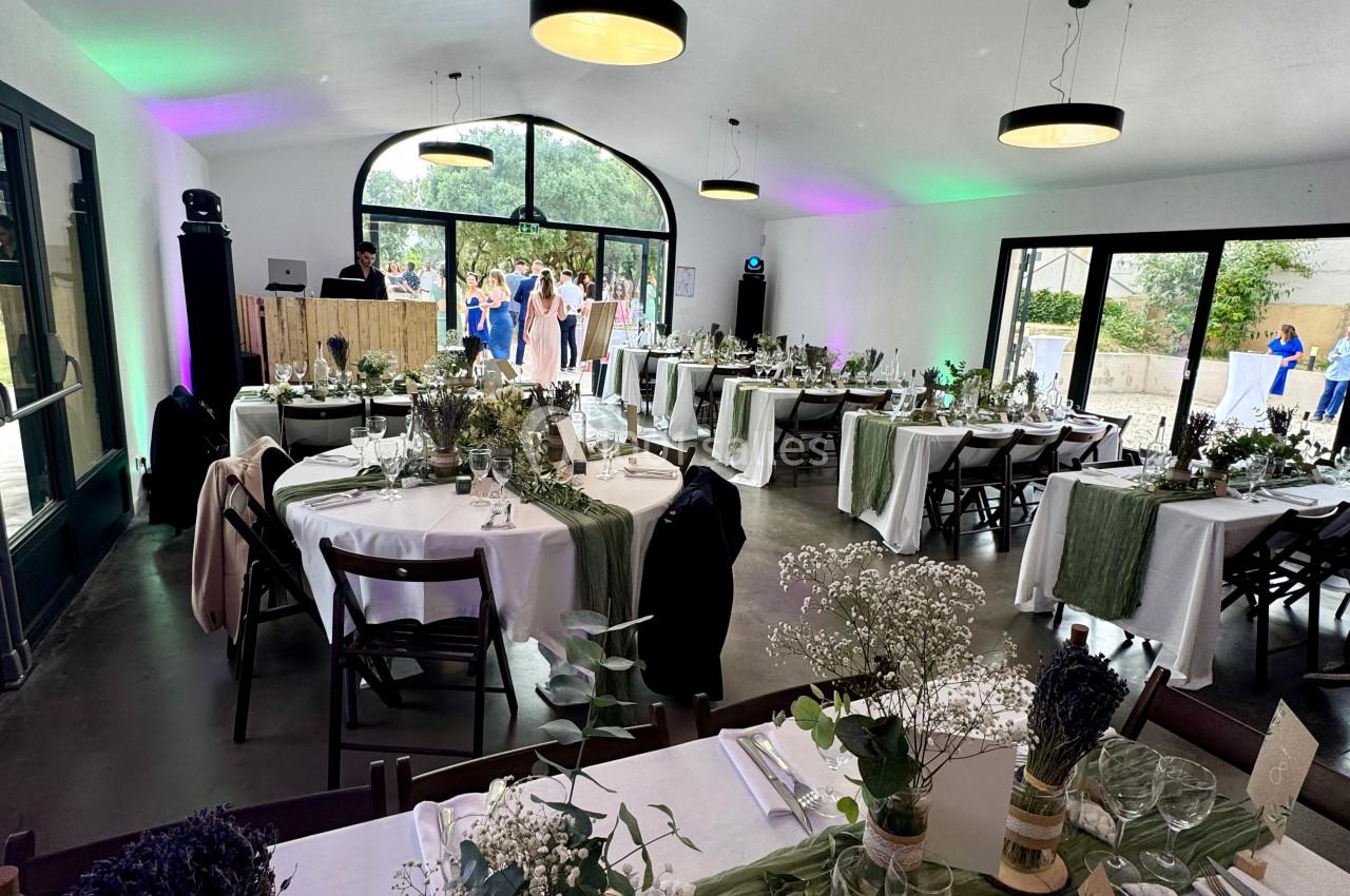 Salle de réception décorée avec des tables dressées, ornées de fleurs et de nappes blanches, éclairée par des suspensions.