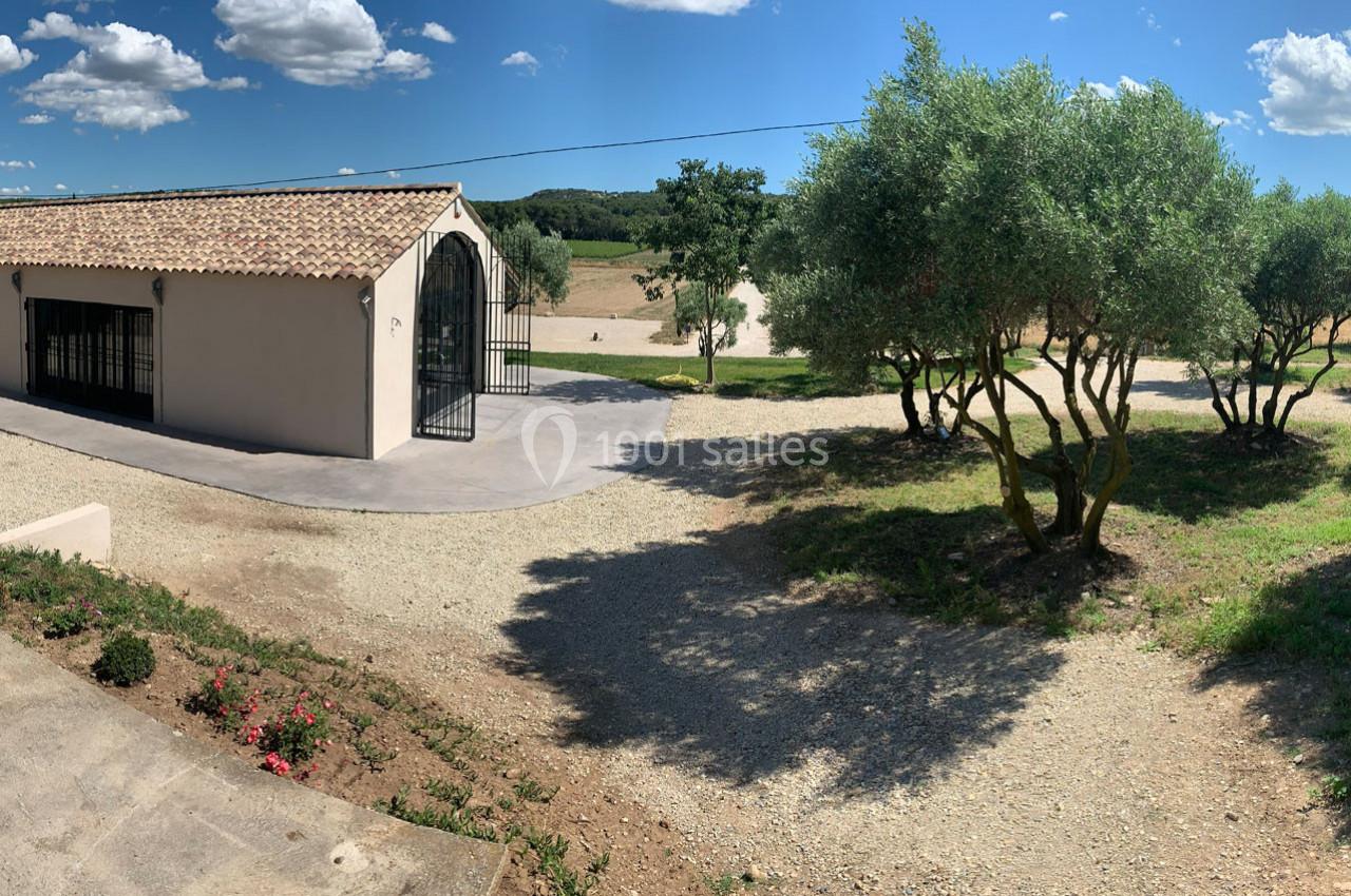 Cour extérieure ensoleillée avec un bâtiment à toit en tuiles, des oliviers et un chemin en gravier.