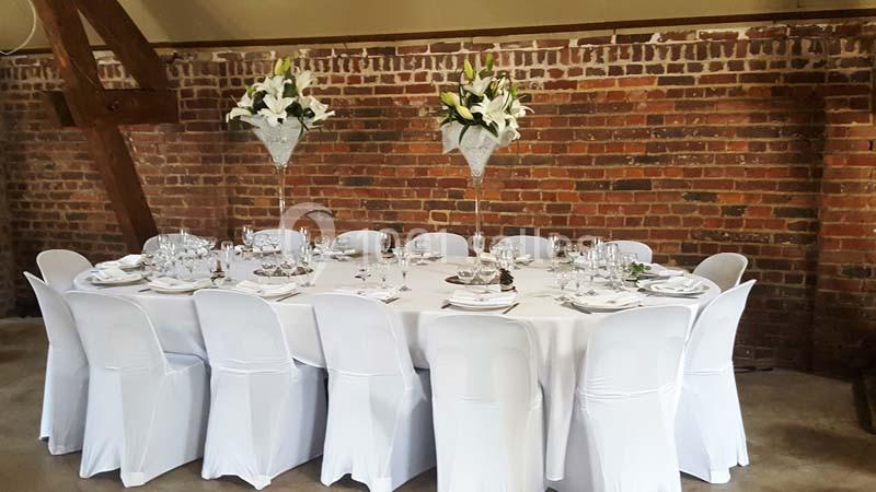 Table ronde dressée pour un événement, nappée de blanc, avec des chaises houssées et des centres de table fleuris.