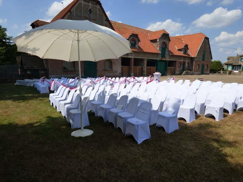 Chaises blanches alignées sous un grand parasol dans une cour extérieure, devant un bâtiment en briques.