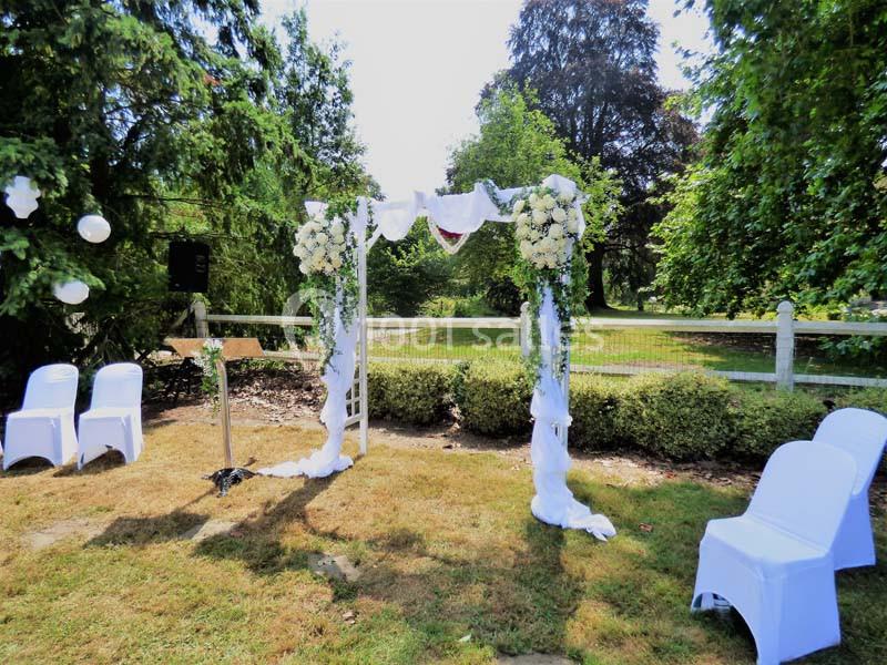 Arche de mariage décorée de fleurs blanches et de tissu, entourée de chaises blanches dans un jardin verdoyant.