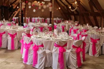 Salle de réception avec tables rondes dressées, décorées de rubans colorés et éclairée par des lustres en bois.