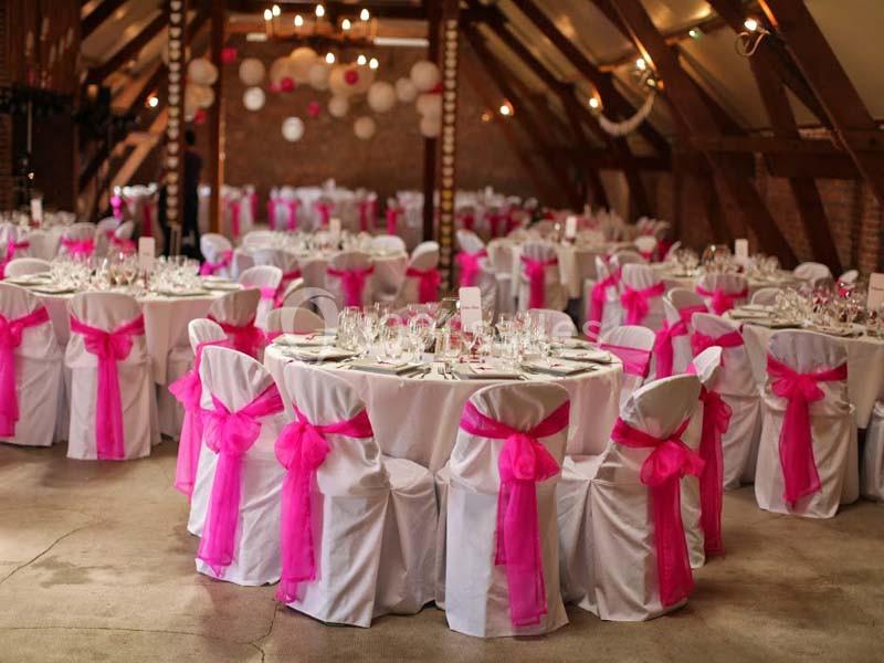Salle décorée pour un événement avec des tables rondes, nappes blanches et chaises ornées de nœuds roses.