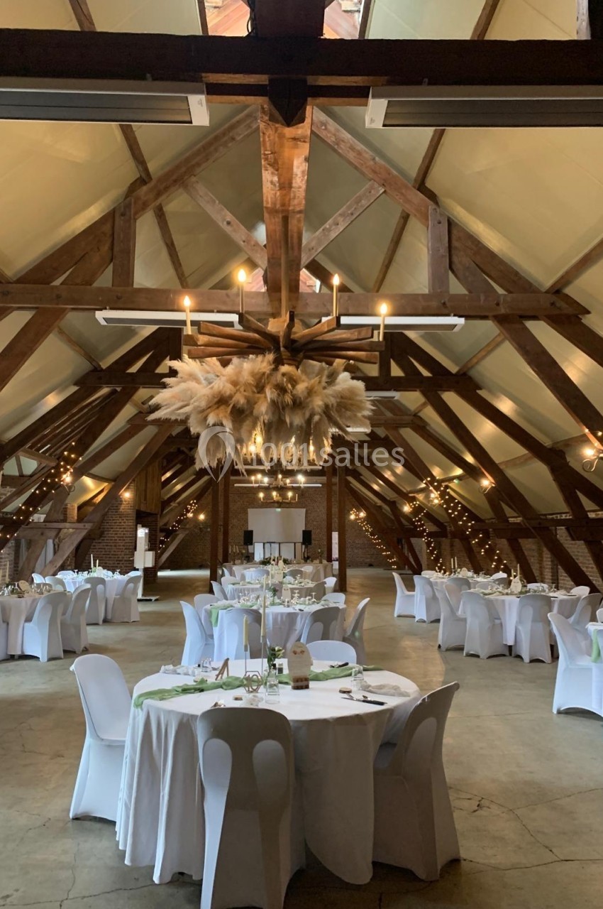 Salle de réception avec charpente en bois, tables rondes dressées et décoration suspendue en pampas.