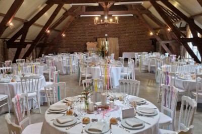 Salle de réception avec tables rondes dressées, décorées de rubans colorés et éclairée par des lustres en bois.