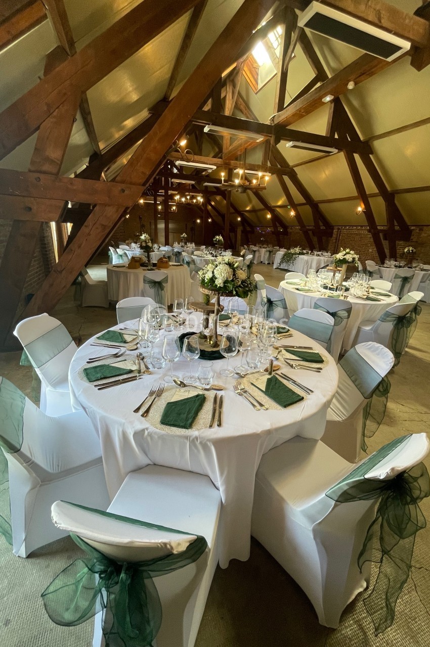 Salle de réception avec poutres apparentes, tables rondes dressées avec nappes blanches et décorations florales.