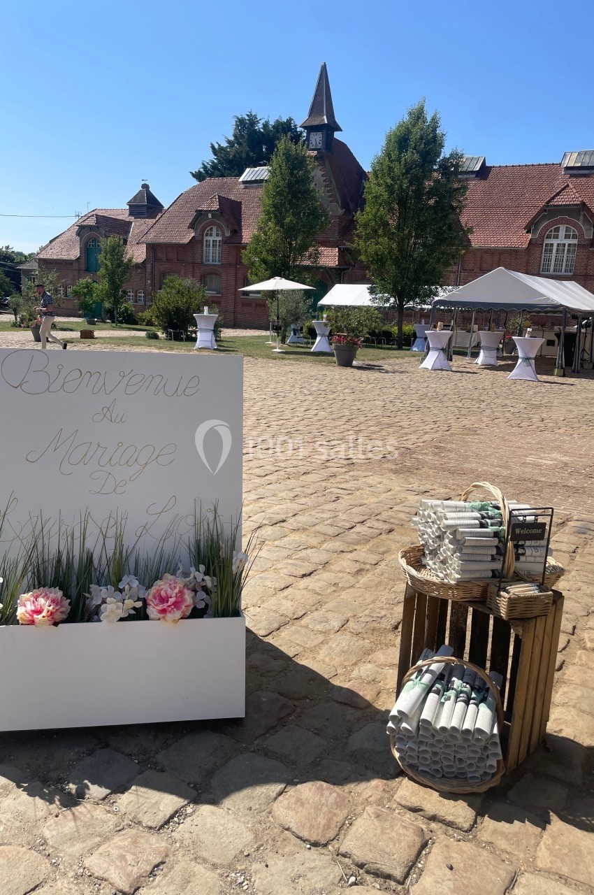 Panneau de bienvenue pour un mariage devant une cour pavée avec tables dressées et bâtiments en briques rouges.