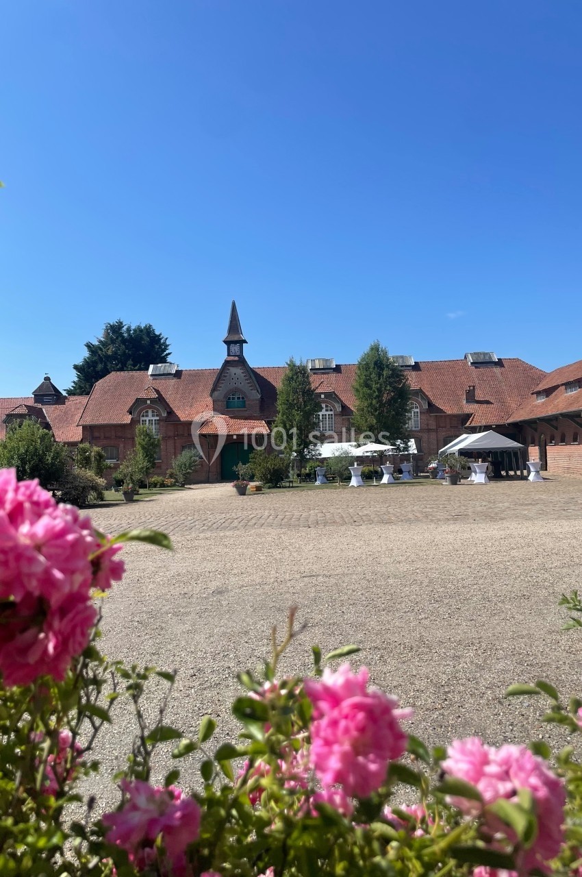Cour pavée devant un bâtiment en briques rouges avec toit en tuiles, fleurs roses au premier plan et ciel bleu.