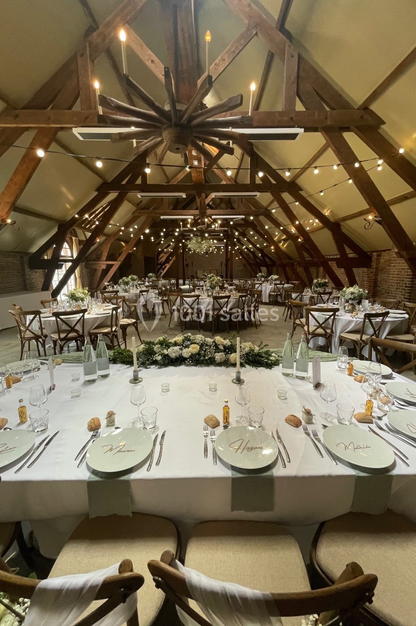 Salle de réception décorée pour un mariage, avec tables dressées, chaises en bois et guirlandes lumineuses suspendues.