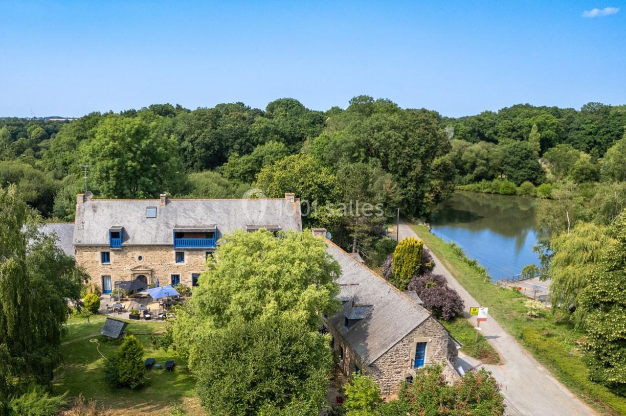 Vue aérienne d'un moulin en pierre entouré de verdure, près d'une rivière et d'un chemin bordé d'arbres.