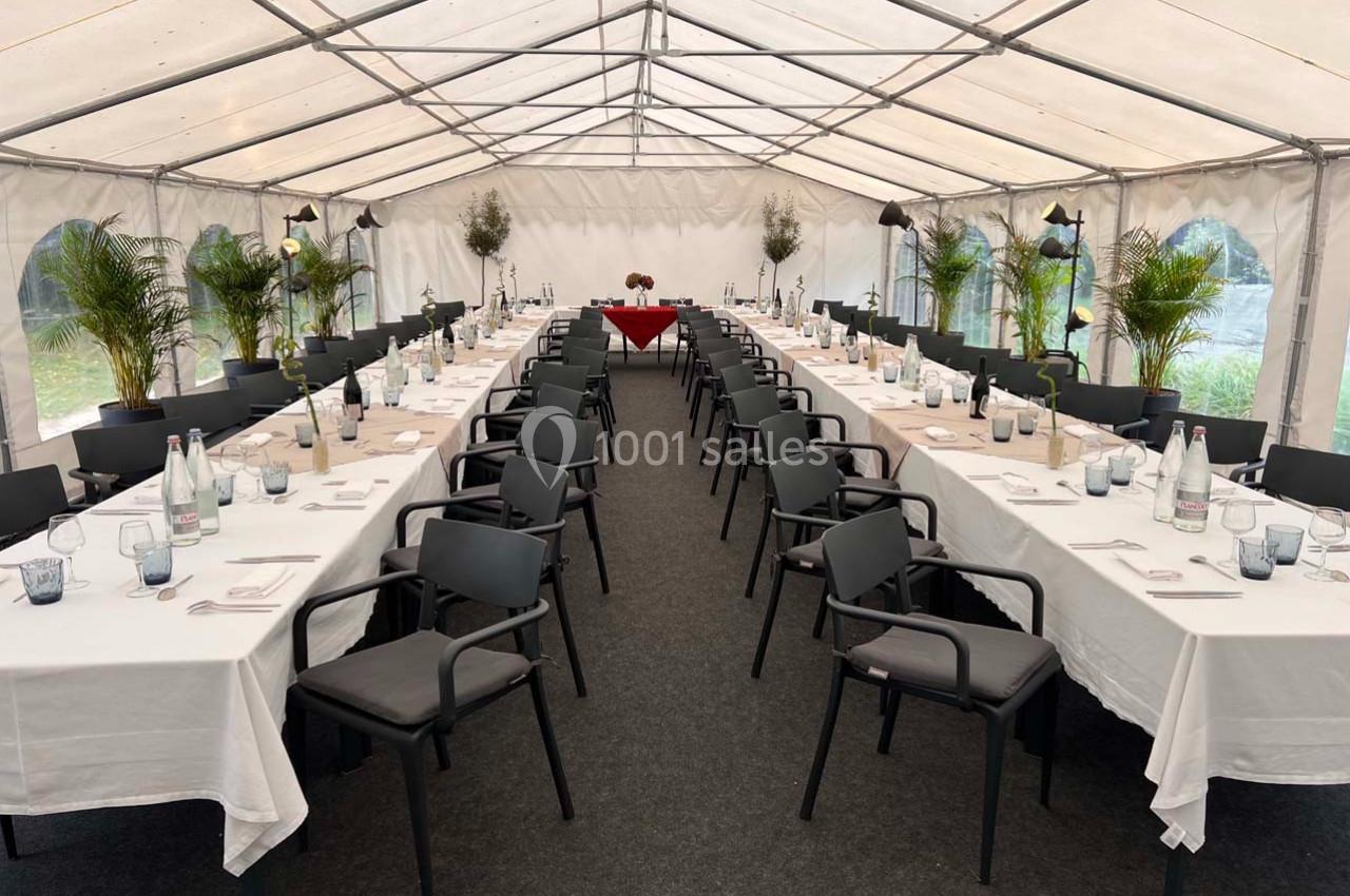 Tables dressées avec nappes blanches et chaises noires alignées dans une grande tente, décorée de plantes vertes.