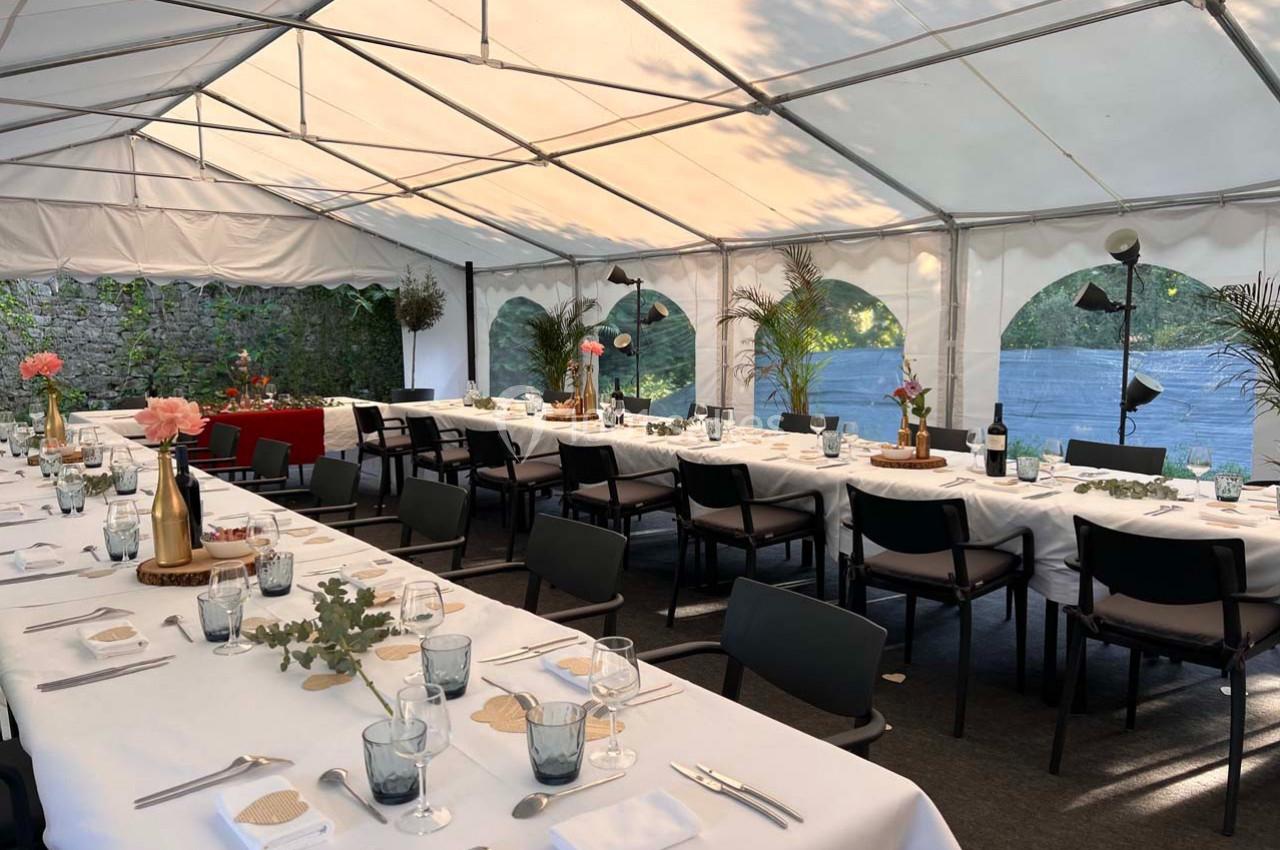 Tables dressées sous une tente blanche, décorées de fleurs et de vaisselle, prêtes pour un événement en extérieur.