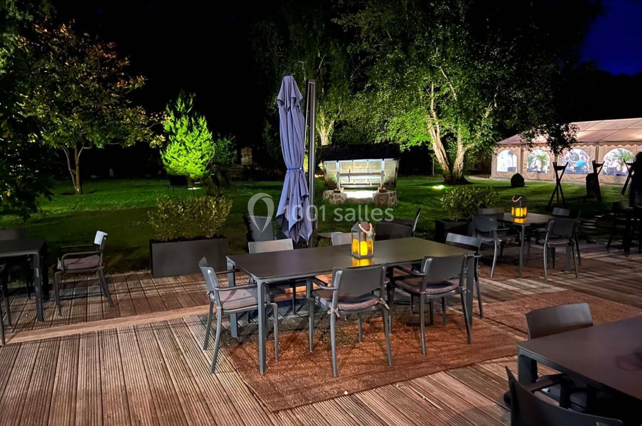 Terrasse en bois éclairée de nuit avec tables, chaises, parasol fermé et vue sur un jardin illuminé en arrière-plan.