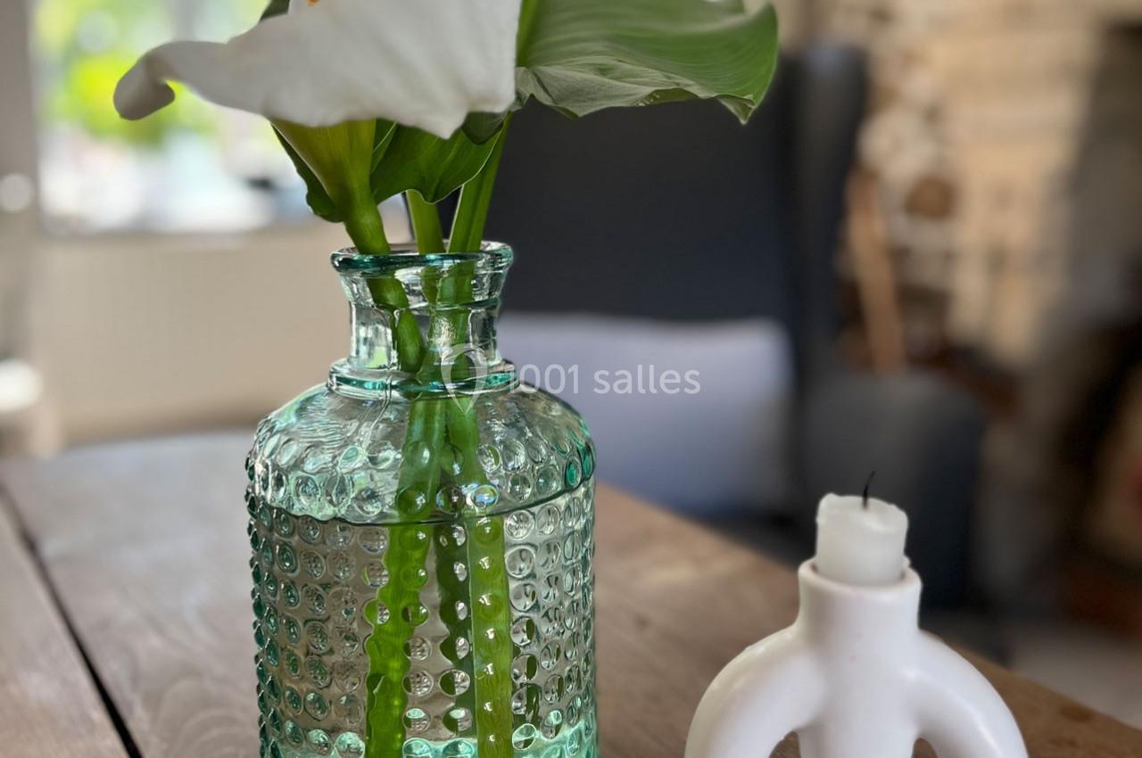 Vase en verre texturé vert contenant des arums blancs, posé sur une table en bois avec un bougeoir blanc à côté.