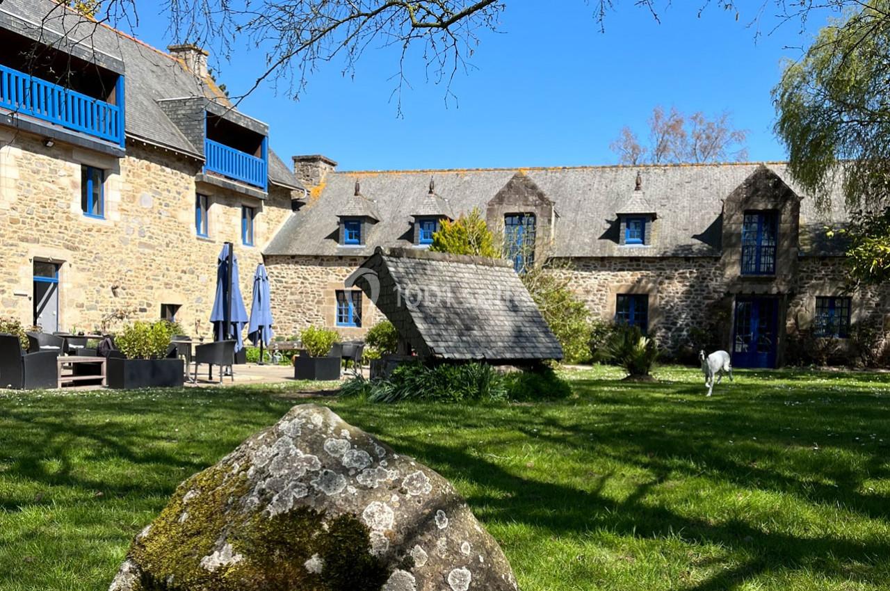Cour verdoyante avec un puits en pierre, entourée de bâtiments en pierre aux volets bleus sous un ciel dégagé.