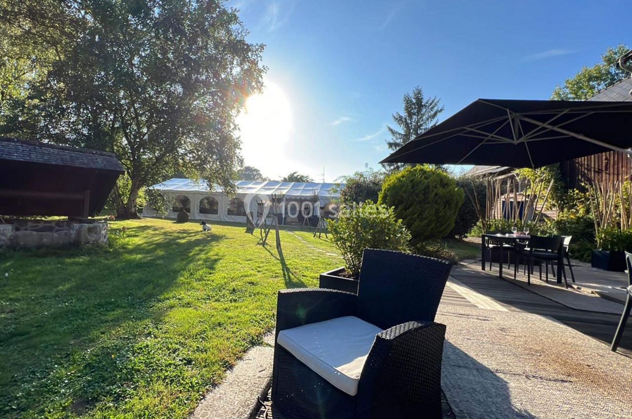 Espace extérieur avec fauteuils, pelouse, arbres et une tente blanche sous un ciel ensoleillé.