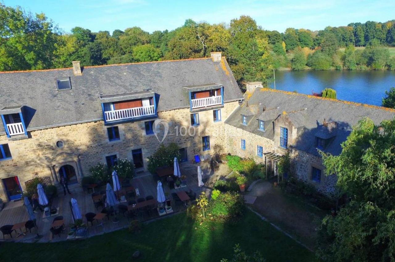 Vue aérienne d'un bâtiment en pierre avec terrasse et tables extérieures, entouré de verdure près d'un plan d'eau.