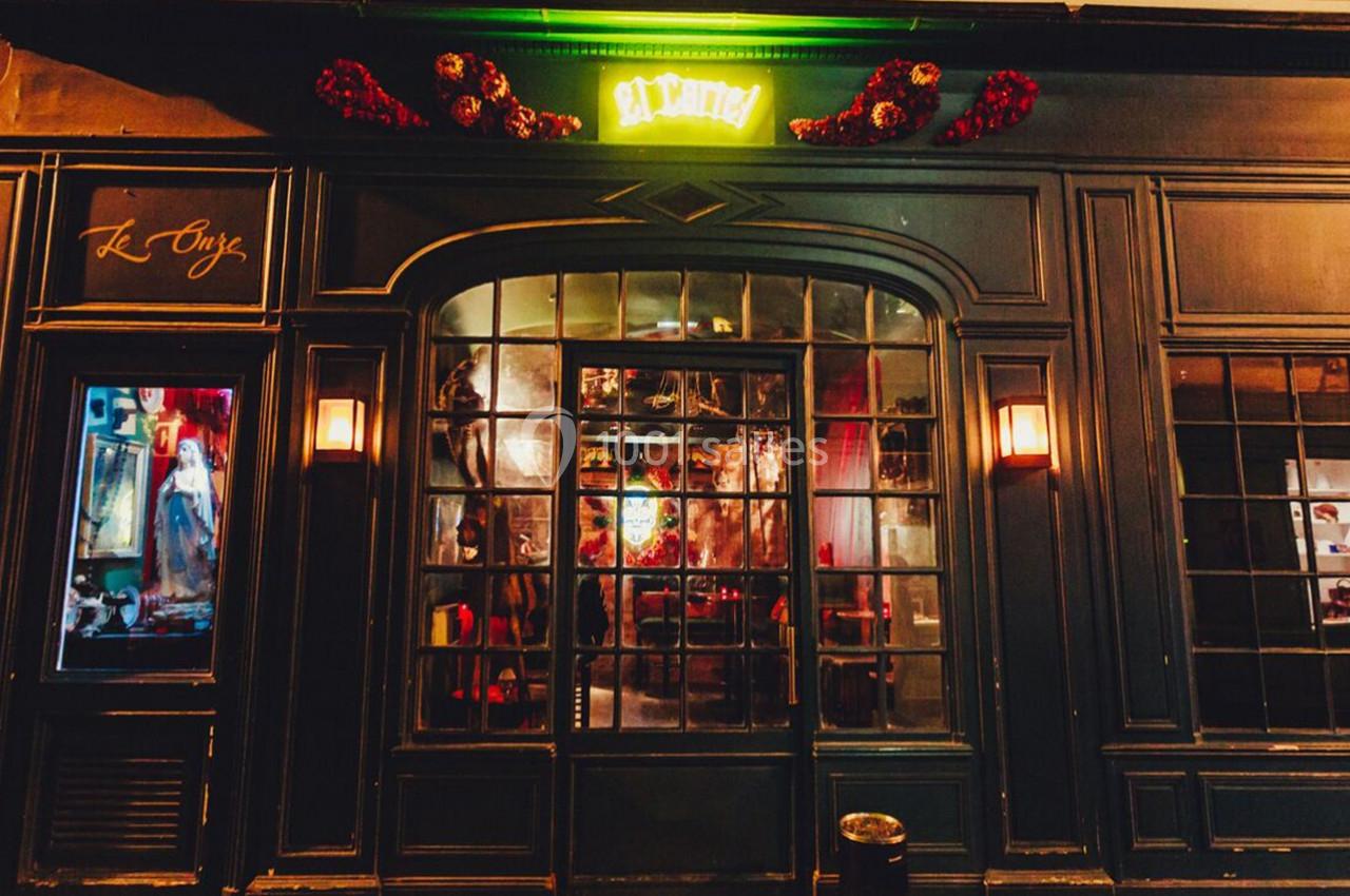 Façade d'une boutique avec vitrine éclairée, enseigne lumineuse verte et décoration florale rouge au-dessus.