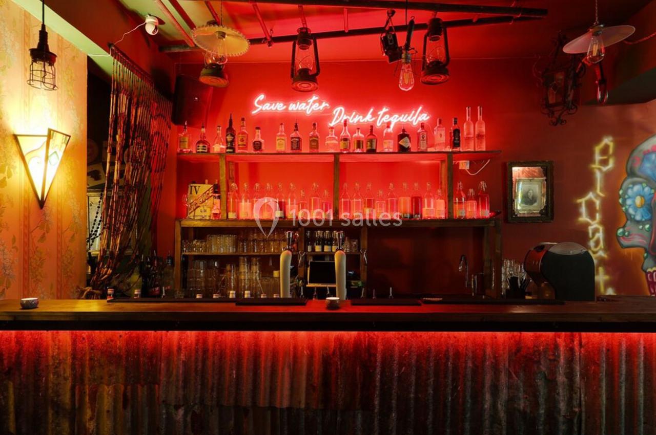 Bar avec éclairage rouge, étagères remplies de bouteilles, néon au mur et décorations murales colorées.