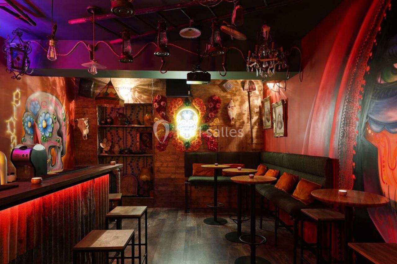 Bar à l'ambiance tamisée avec décorations murales colorées, tables hautes et éclairage rouge chaleureux.