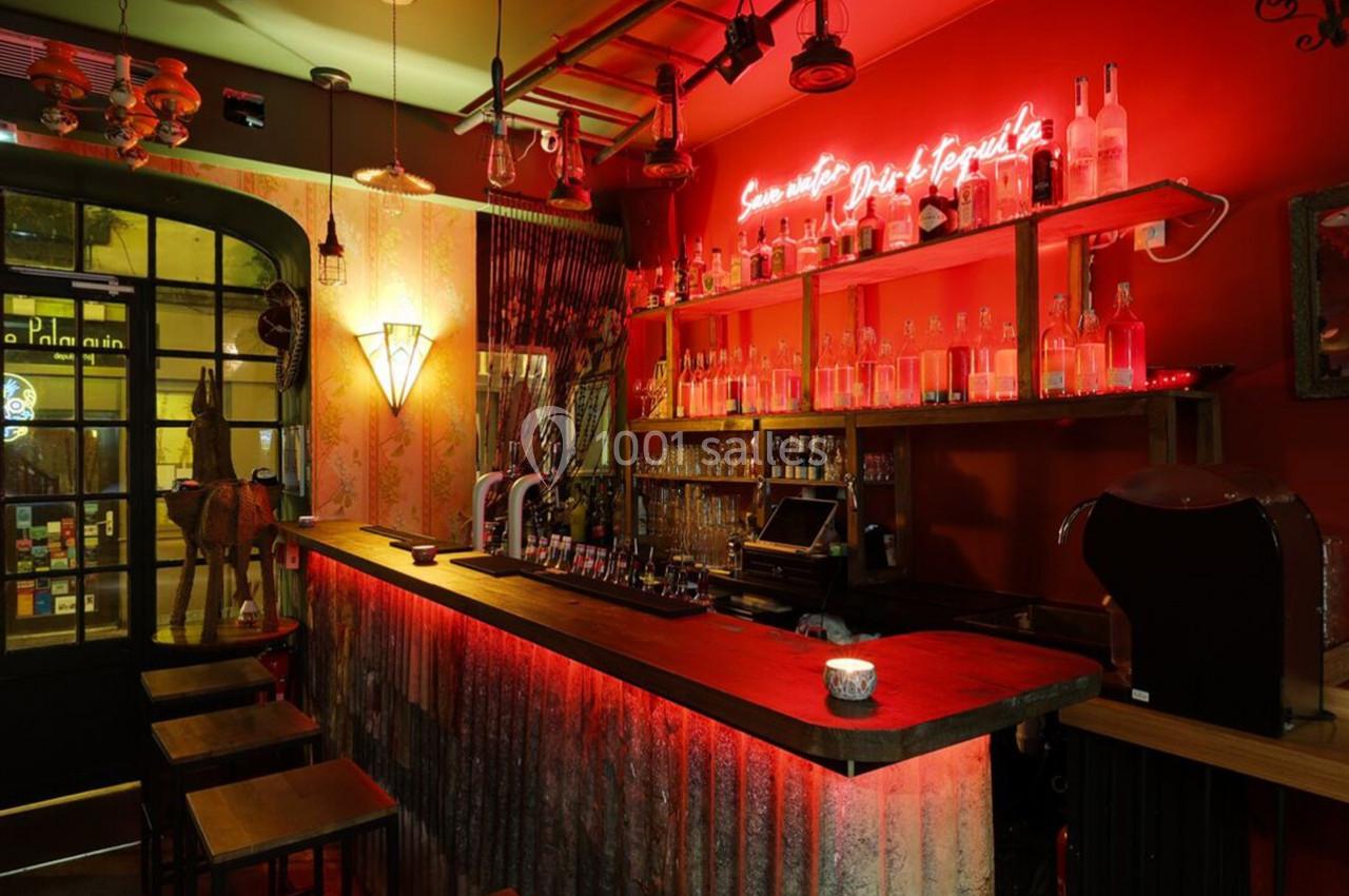 Bar à l'ambiance chaleureuse avec éclairage rouge, comptoir en bois, étagères de bouteilles et néon lumineux au mur.