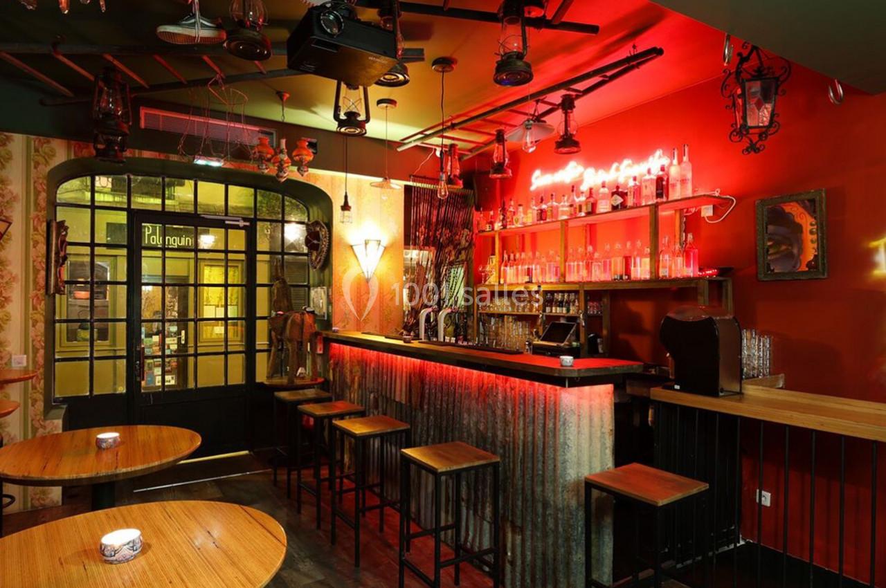 Intérieur d'un bar avec comptoir en métal, éclairage rouge, tables en bois et décorations murales éclectiques.