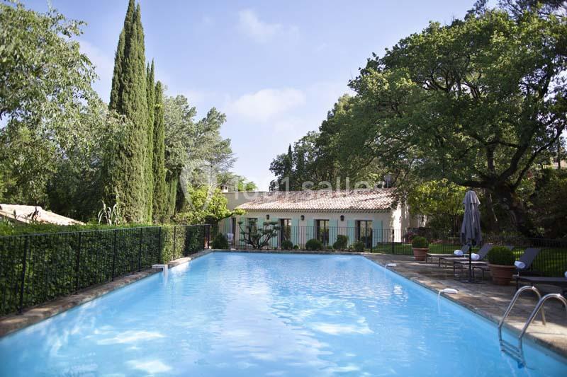 Piscine extérieure entourée de végétation, avec une maison à toit en tuiles en arrière-plan sous un ciel dégagé.