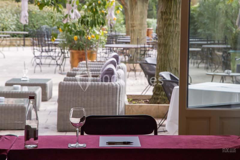 Table avec nappe rouge, verre, carafe et papier, donnant sur une terrasse avec mobilier de jardin et arbres.