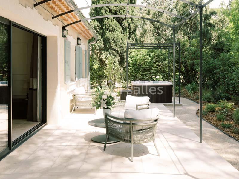 Terrasse avec mobilier de jardin, pergola métallique, jacuzzi en arrière-plan et végétation environnante.
