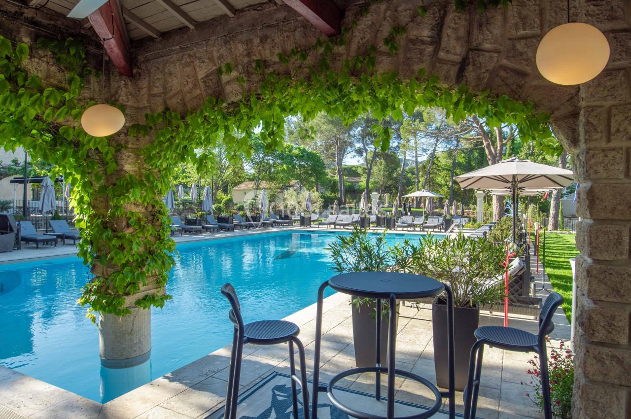 Terrasse ombragée avec table et chaises donnant sur une piscine entourée de transats et de végétation.