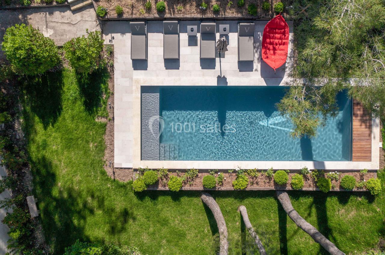 Vue aérienne d'une piscine rectangulaire entourée de chaises longues, d'un parasol rouge et de verdure.