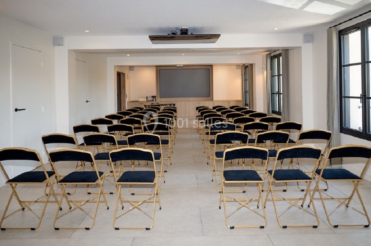 Salle de réunion lumineuse avec des rangées de chaises pliantes noires face à un écran de projection.