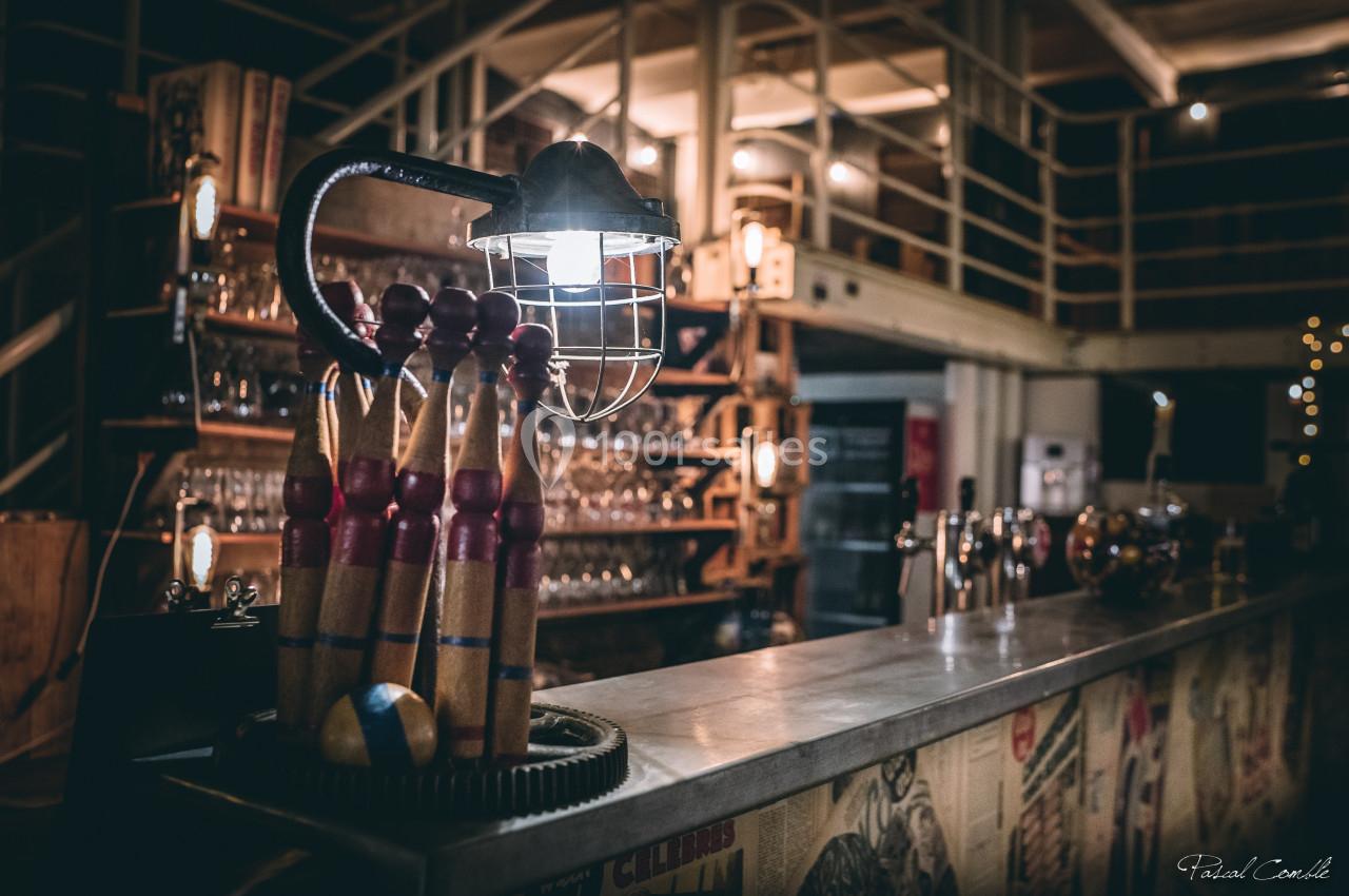 Lampe industrielle éclairant un comptoir en métal, décoré de quilles en bois, dans un bar à l'ambiance rétro.