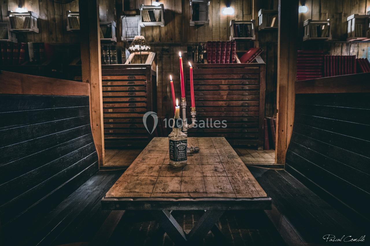 Table en bois avec une bouteille servant de chandelier à trois bougies rouges, dans une pièce rustique avec des étagères et…
