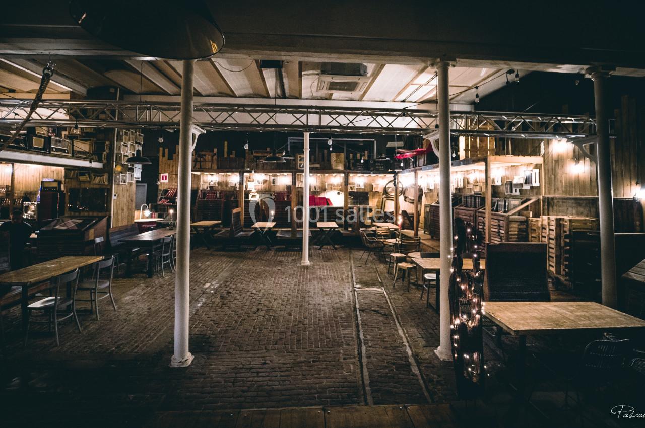 Salle intérieure avec tables et chaises en bois, éclairée par des lumières chaleureuses, ambiance rustique et industrielle.