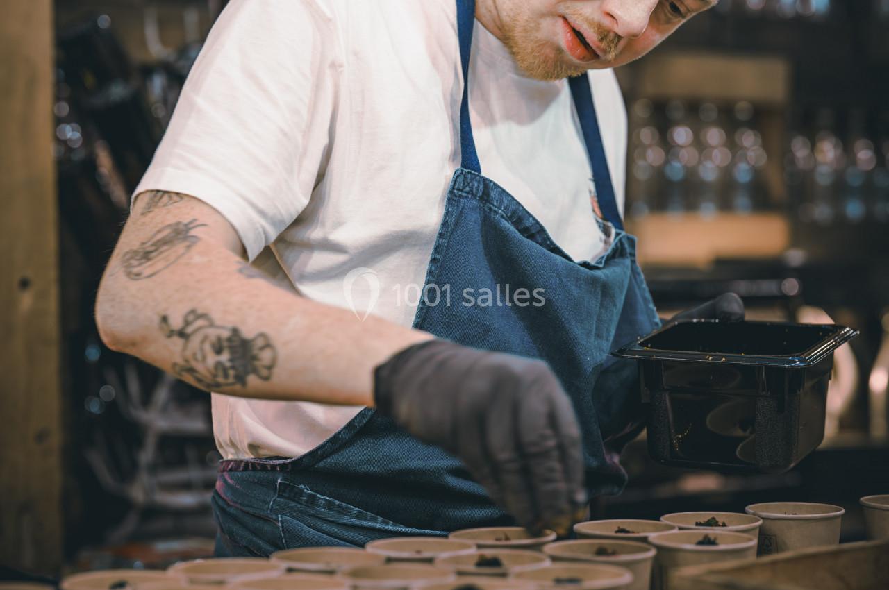 Homme en tablier bleu préparant des plats dans des contenants individuels sur un plan de travail en bois.
