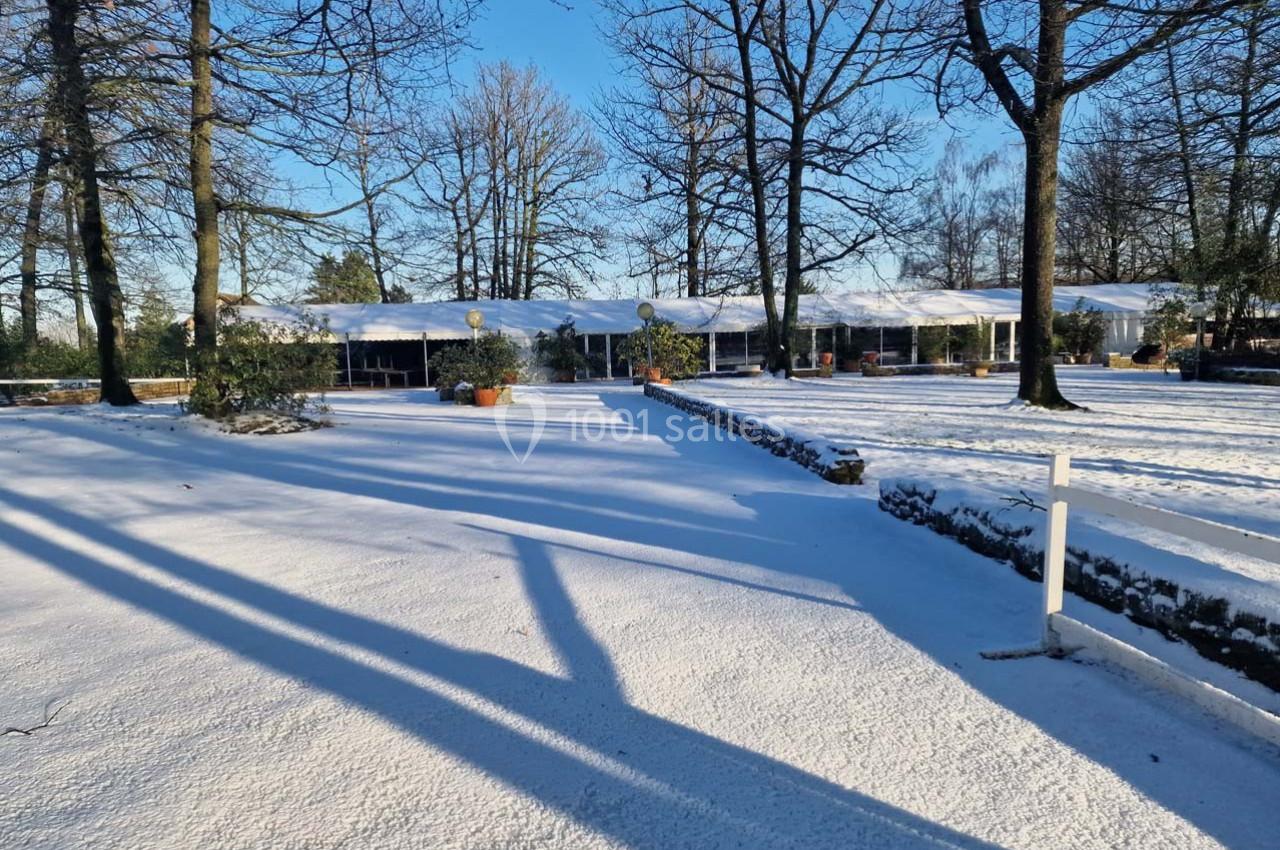 Paysage hivernal avec un bâtiment bas entouré d'arbres, sol recouvert de neige et ombres longues sous un ciel bleu.