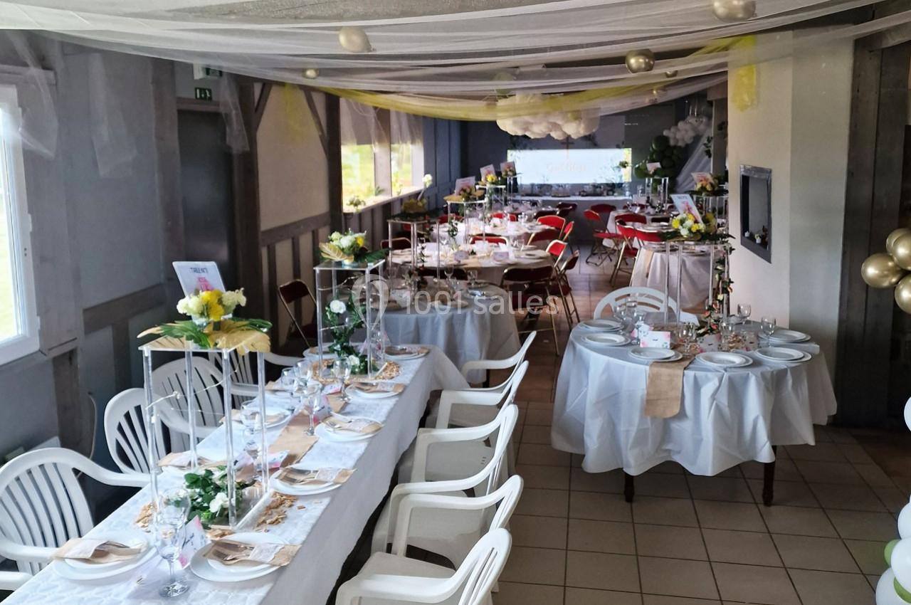 Salle décorée pour un événement avec tables dressées, nappes blanches, fleurs et chaises en plastique.
