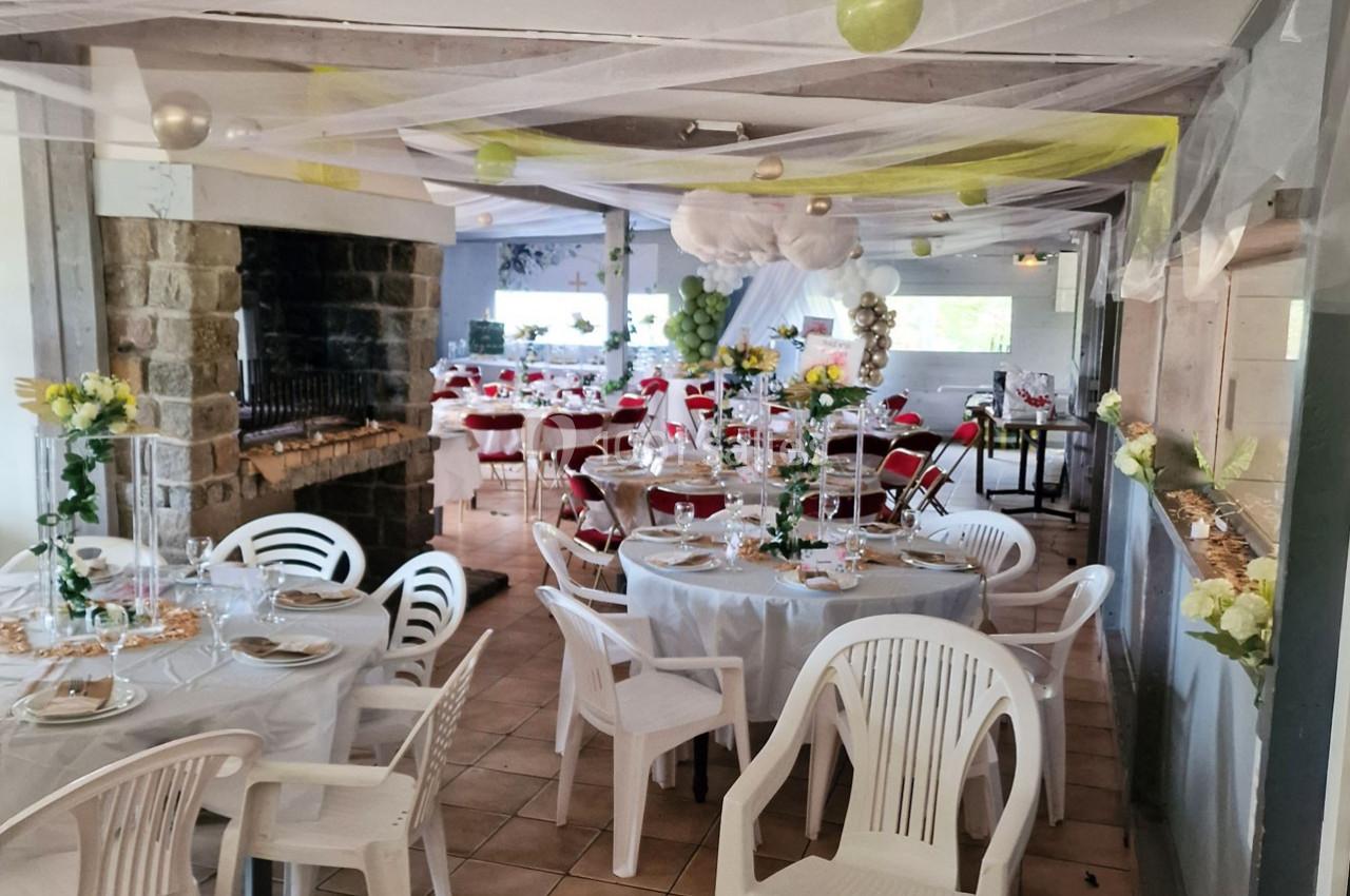 Salle décorée pour un événement avec tables rondes dressées, chaises blanches et ornements suspendus.