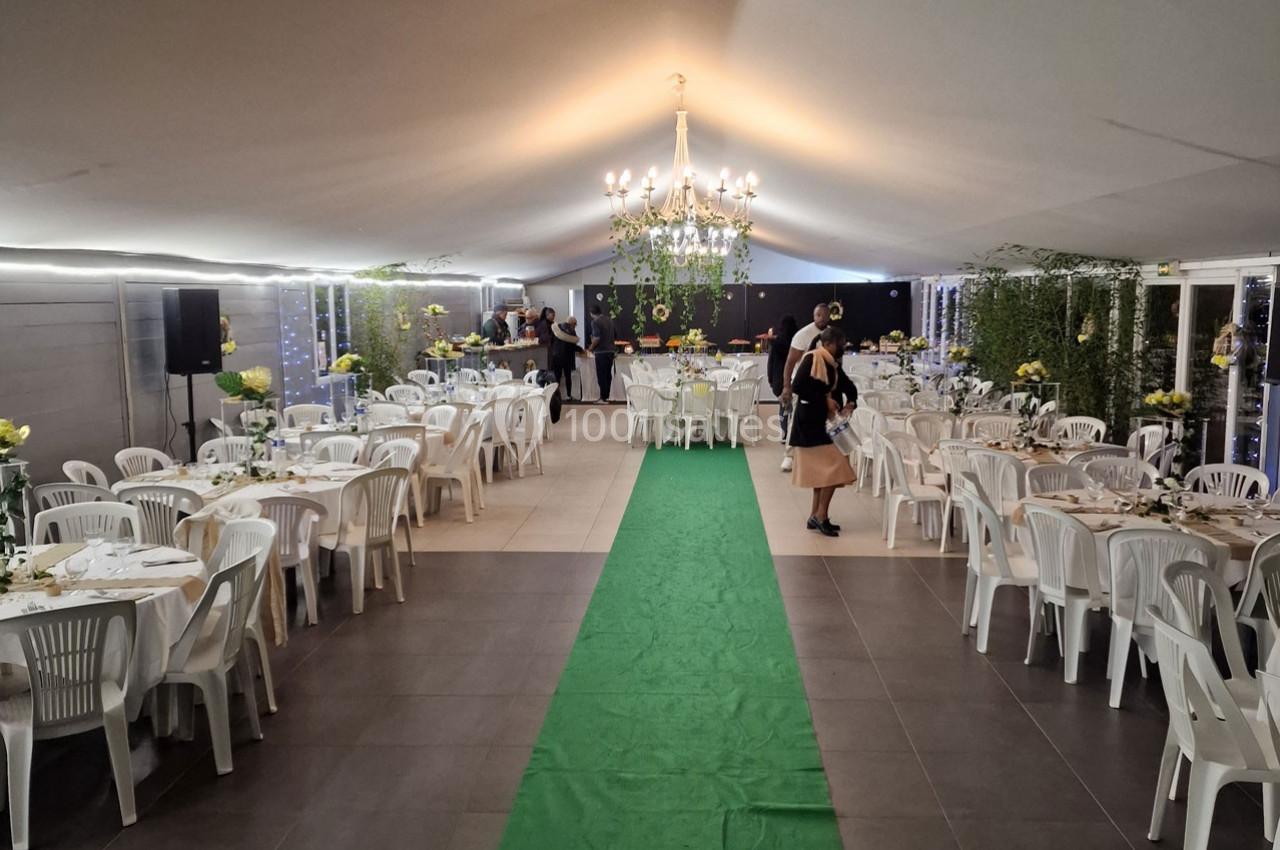 Salle de réception décorée avec des tables rondes, des chaises blanches, un tapis vert et un lustre central.
