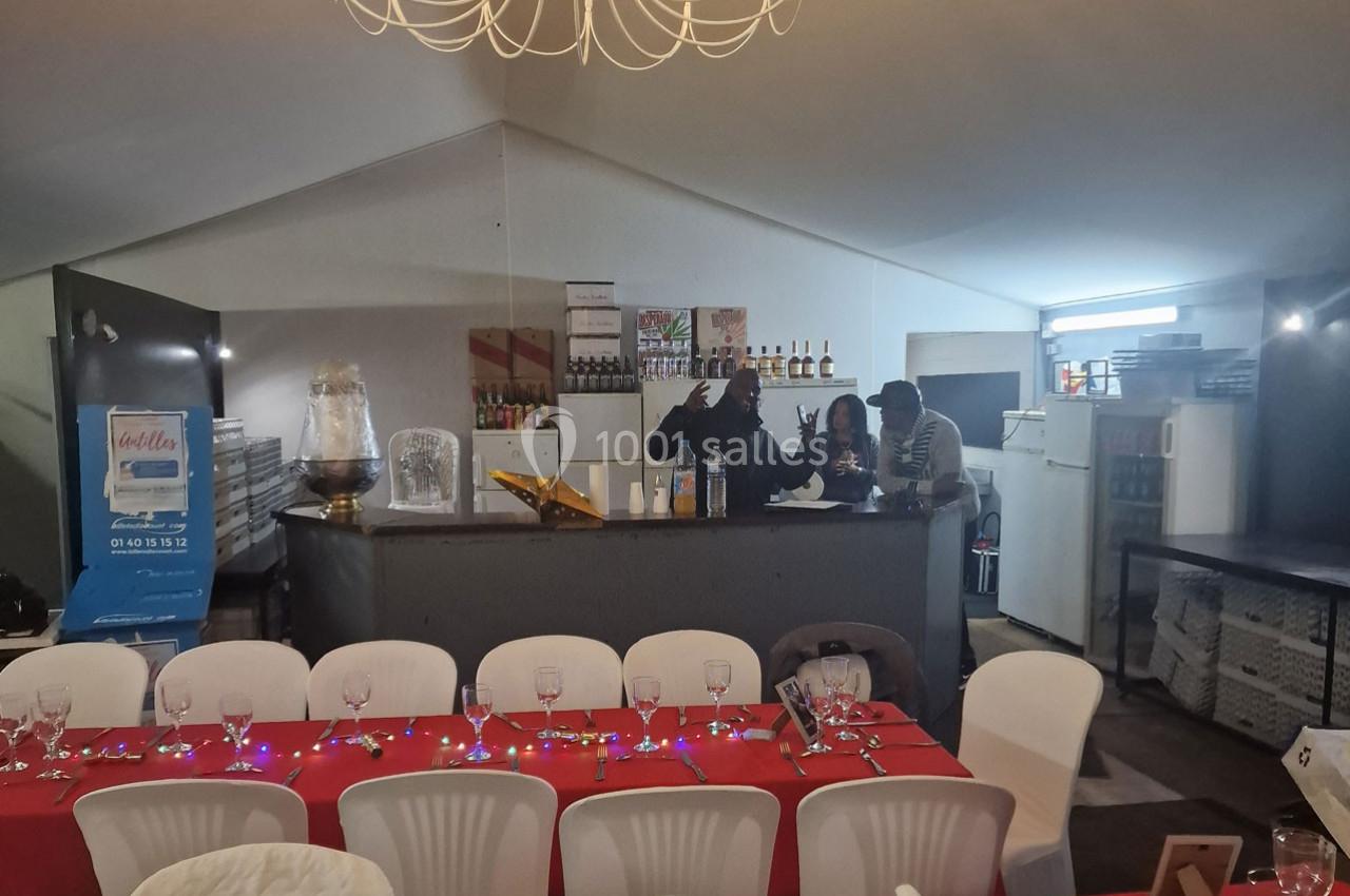 Salle de réception avec tables dressées en rouge et blanc, bar en arrière-plan avec trois personnes discutant.